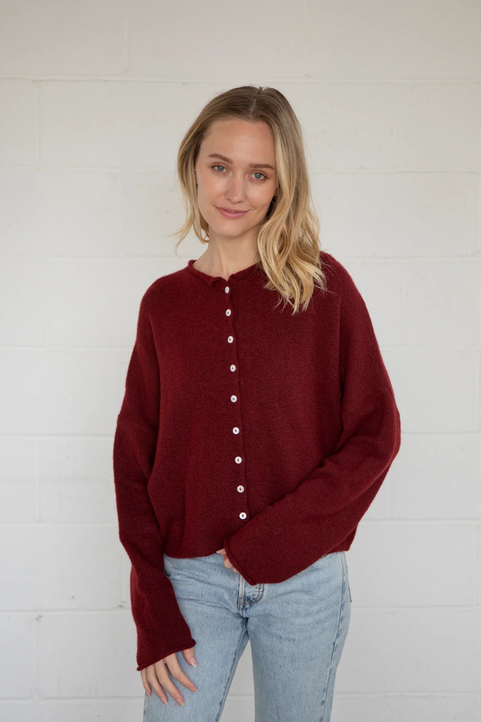 Woman wearing a burgundy button-up cardigan sweter and light blue jeans against a white brick wall.