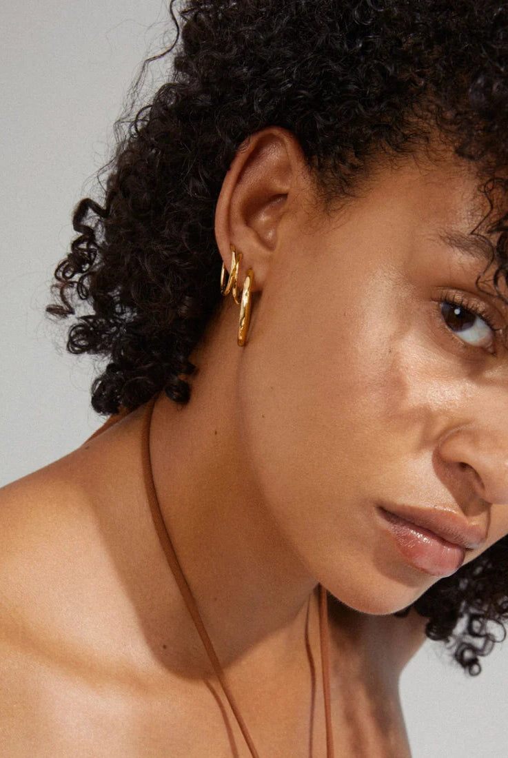 Close-up of a person with curly hair wearing gold hoop earrings against a neutral background