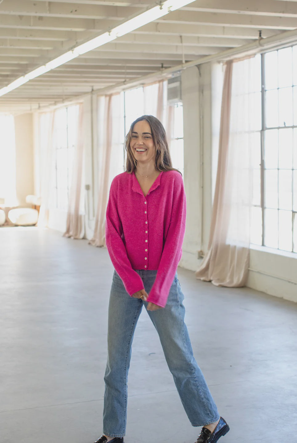 Person wearing a bright pink cardigan and blue jeans standing in a room with large windows and white walls.