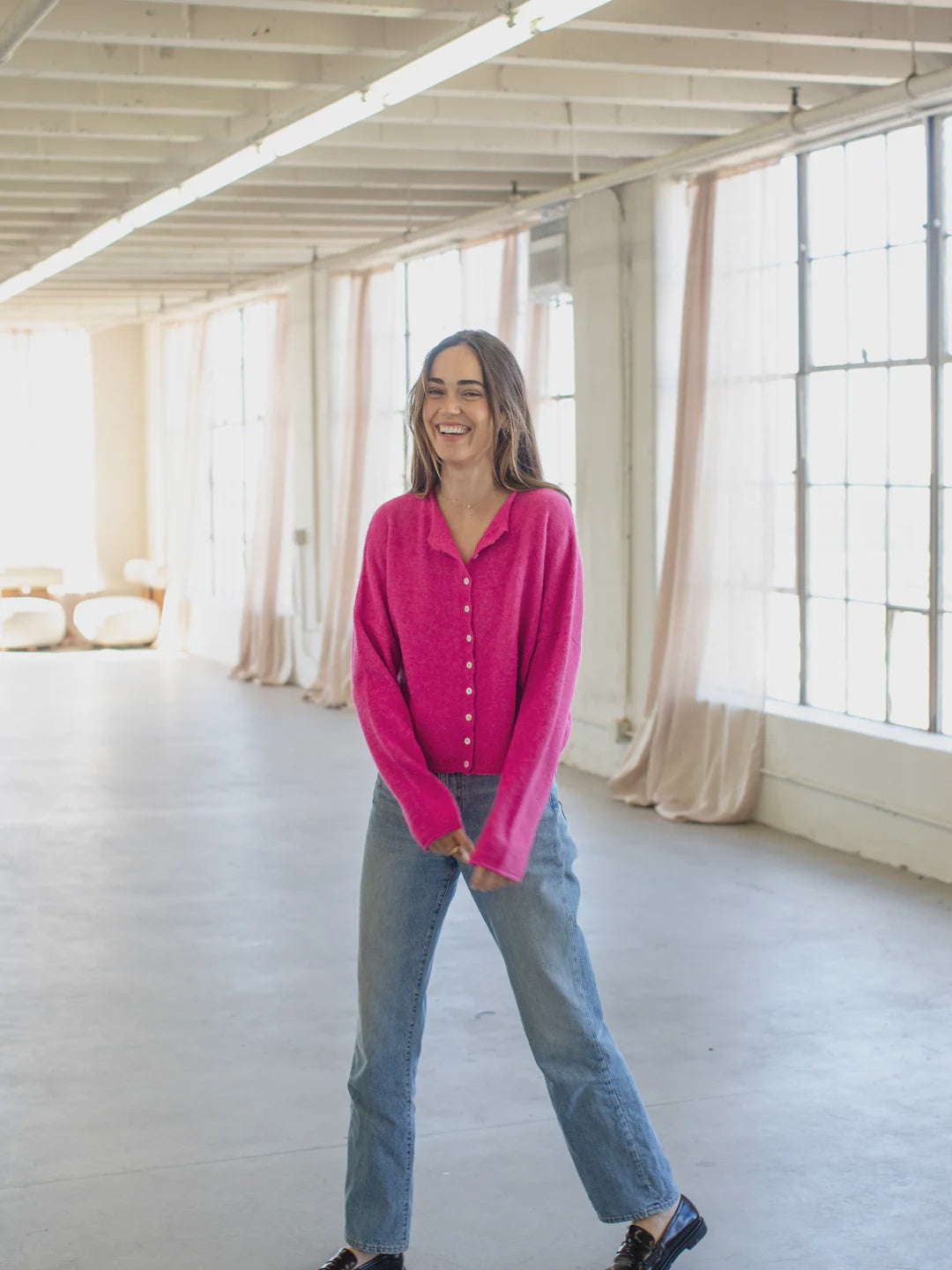 Person wearing a bright pink cardigan and blue jeans standing in a room with large windows and white walls.