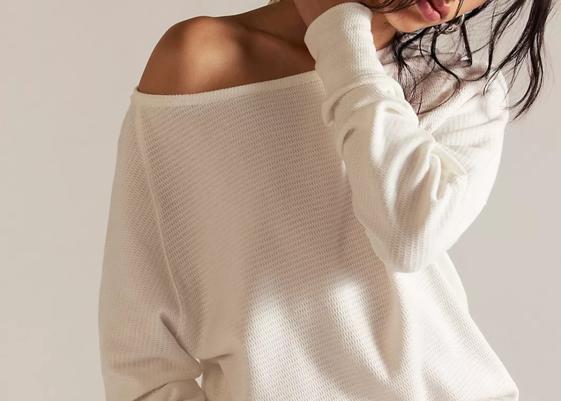 Woman wearing a white off-shoulder sweater and light-colored shorts against a plain background
