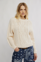 Woman wearing a cream cable knit sweater and blue patterned pants on a plain background