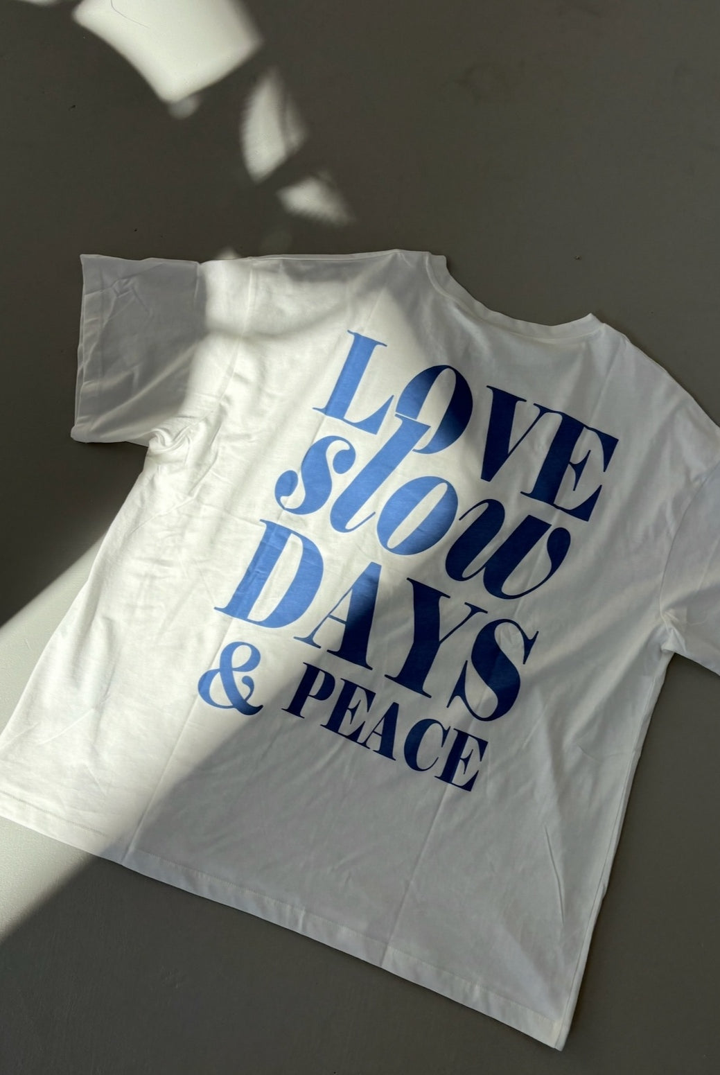 White t-shirt with blue text 'LOVE slow DAYS & PEACE' on a dark surface