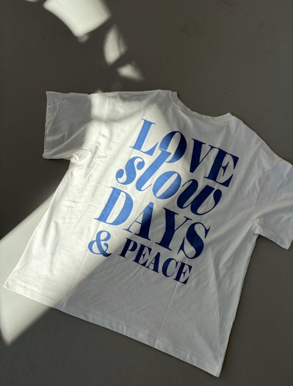 White t-shirt with blue text 'LOVE slow DAYS & PEACE' on a dark surface