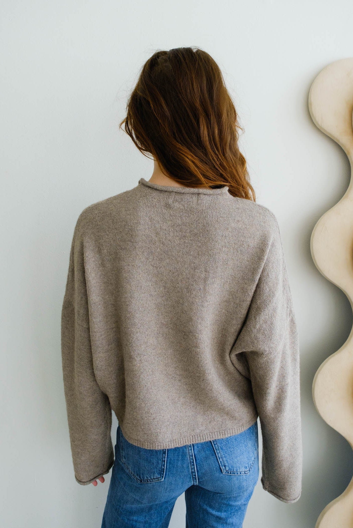 Person wearing a beige sweater and blue jeans against a white wall with decorative elements.