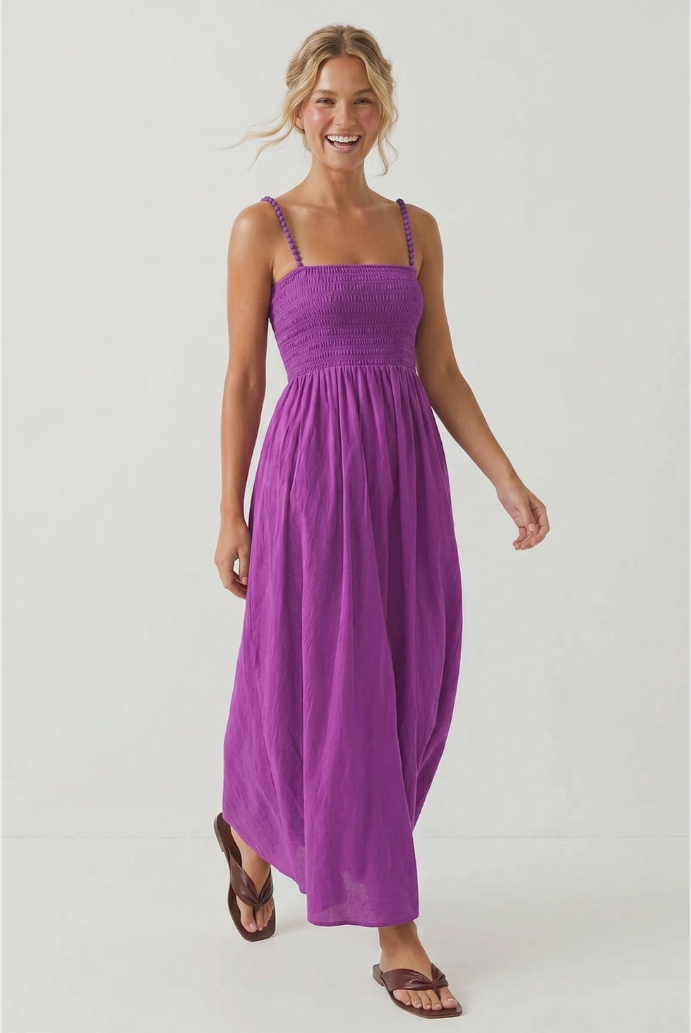 Purple maxi dress featuring a fitted smocked bodice, square neckline, adjustable pom pom shoulder straps, a flowing full-length skirt, and side pockets