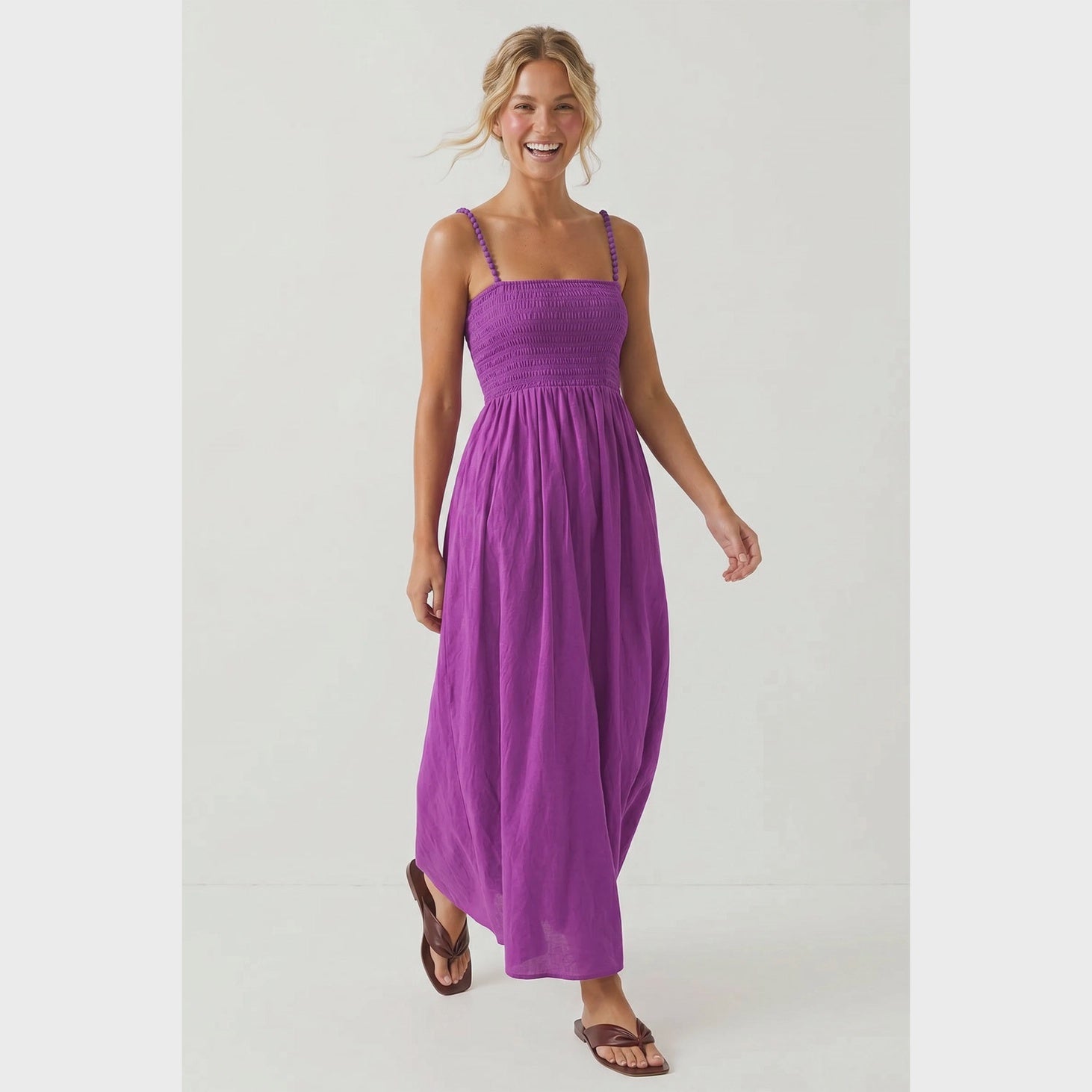 Purple maxi dress featuring a fitted smocked bodice, square neckline, adjustable pom pom shoulder straps, a flowing full-length skirt, and side pockets