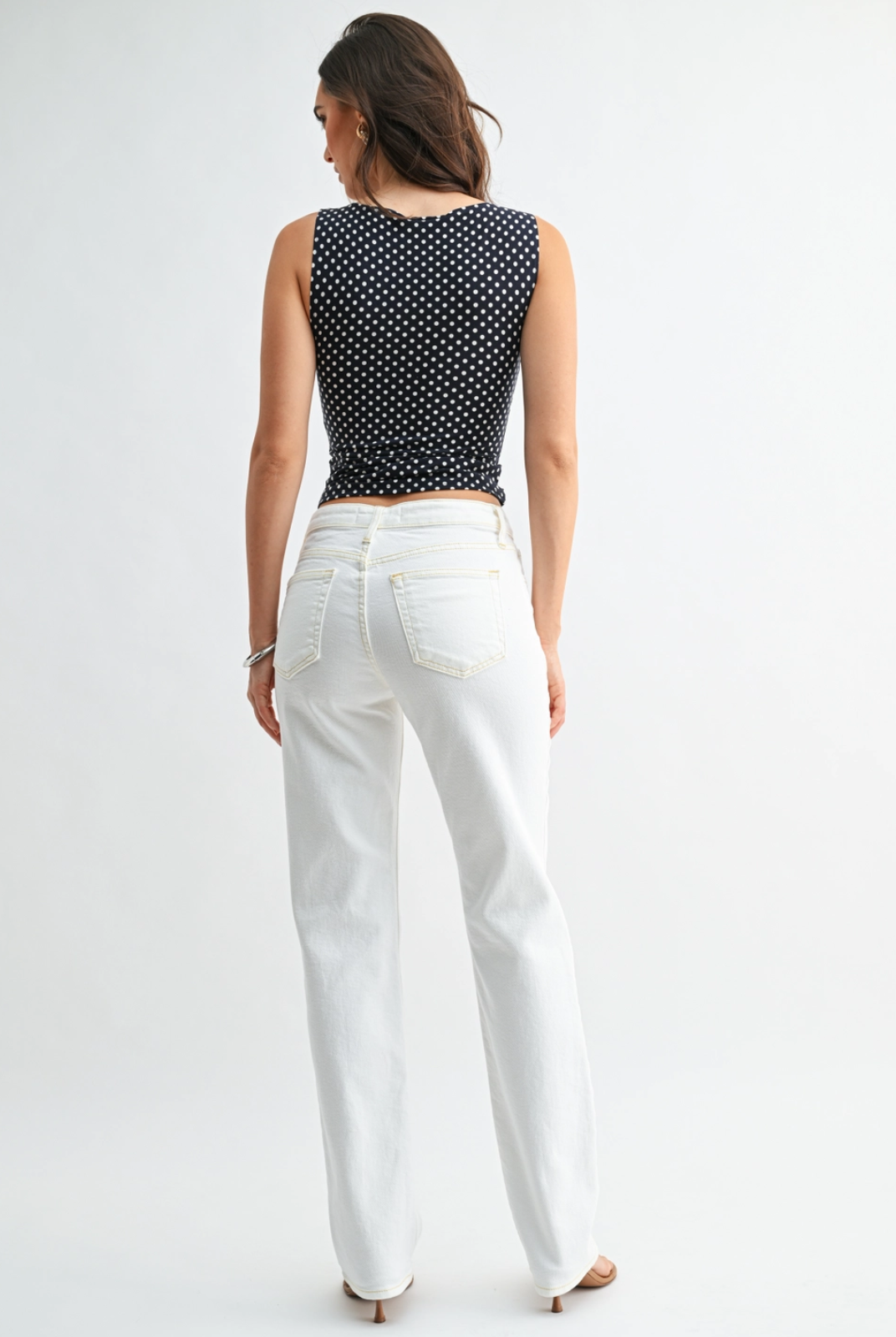 Woman wearing a black and white polka dot top and white pants on a white background. Lee & Me Boutique