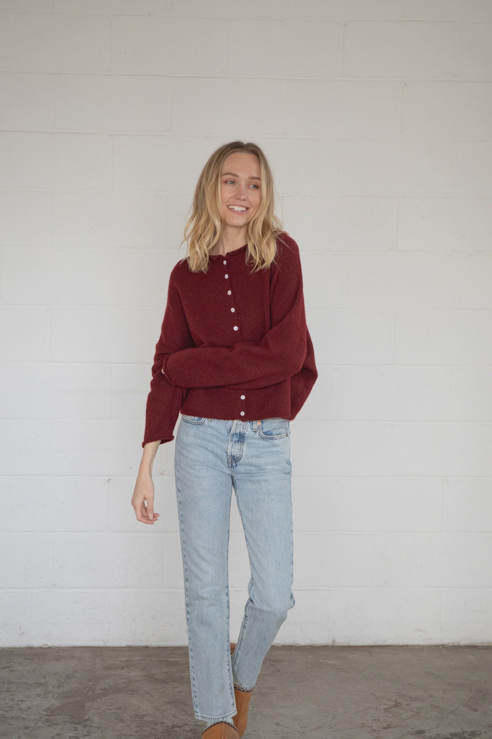 Woman wearing a burgundy sweater and light blue jeans against a white wall.