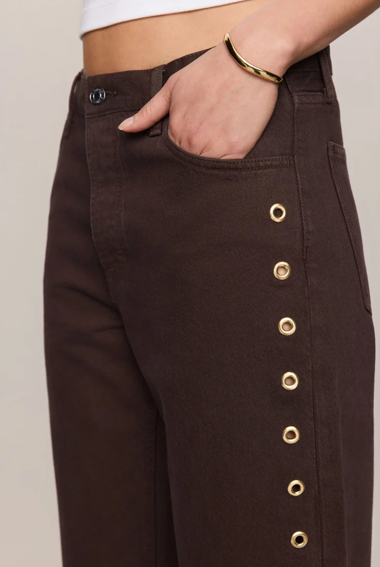 Rich brown mid-rise denim jeans featuring a relaxed bowed-leg silhouette, ankle-length hem, gold grommet details, and subtle side openings.