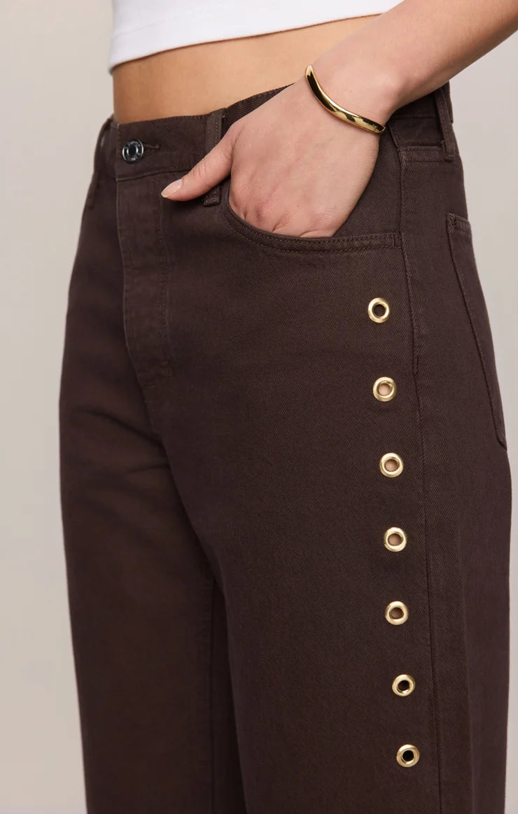 Rich brown mid-rise denim jeans featuring a relaxed bowed-leg silhouette, ankle-length hem, gold grommet details, and subtle side openings.