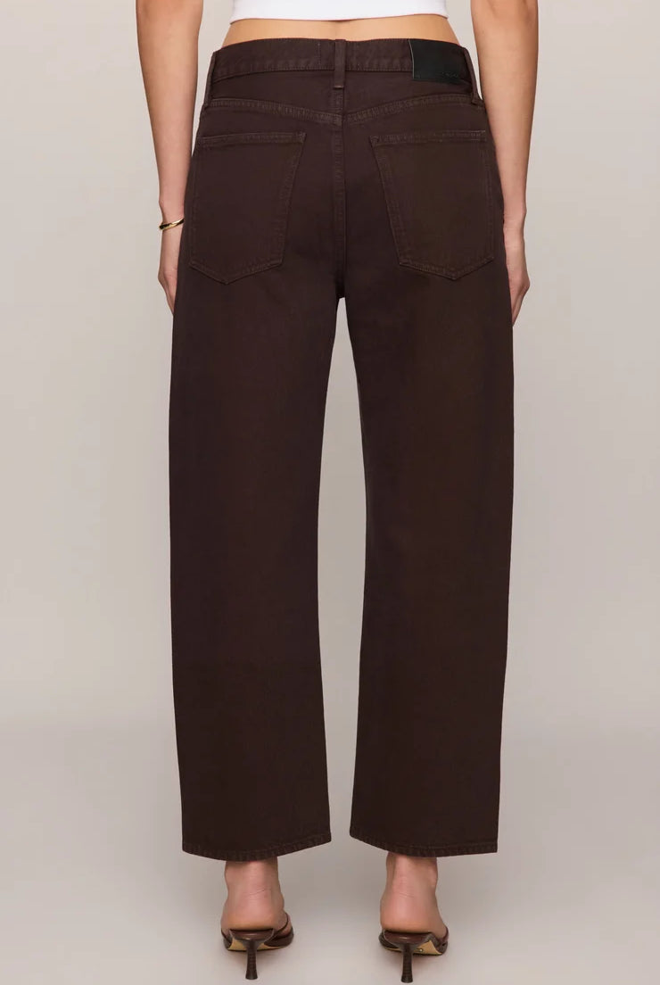 Rich brown mid-rise denim jeans featuring a relaxed bowed-leg silhouette, ankle-length hem, gold grommet details, and subtle side openings.