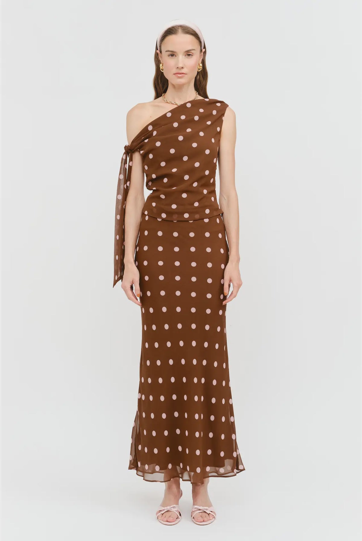 Celestine Off the Shoulder Top in brown featuring soft pink polka dots, an asymmetrical one-shoulder neckline, and a draped tie detail along the arm.