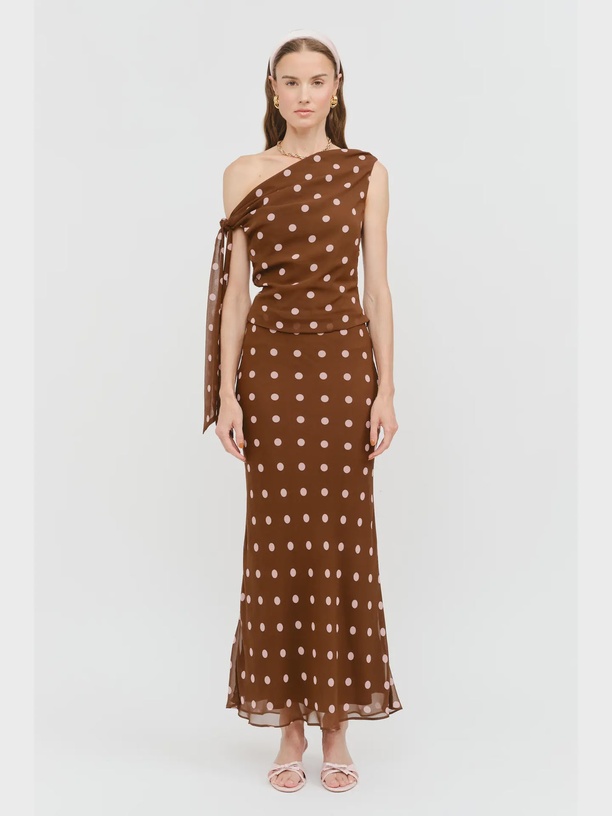Celestine Off the Shoulder Top in brown featuring soft pink polka dots, an asymmetrical one-shoulder neckline, and a draped tie detail along the arm.