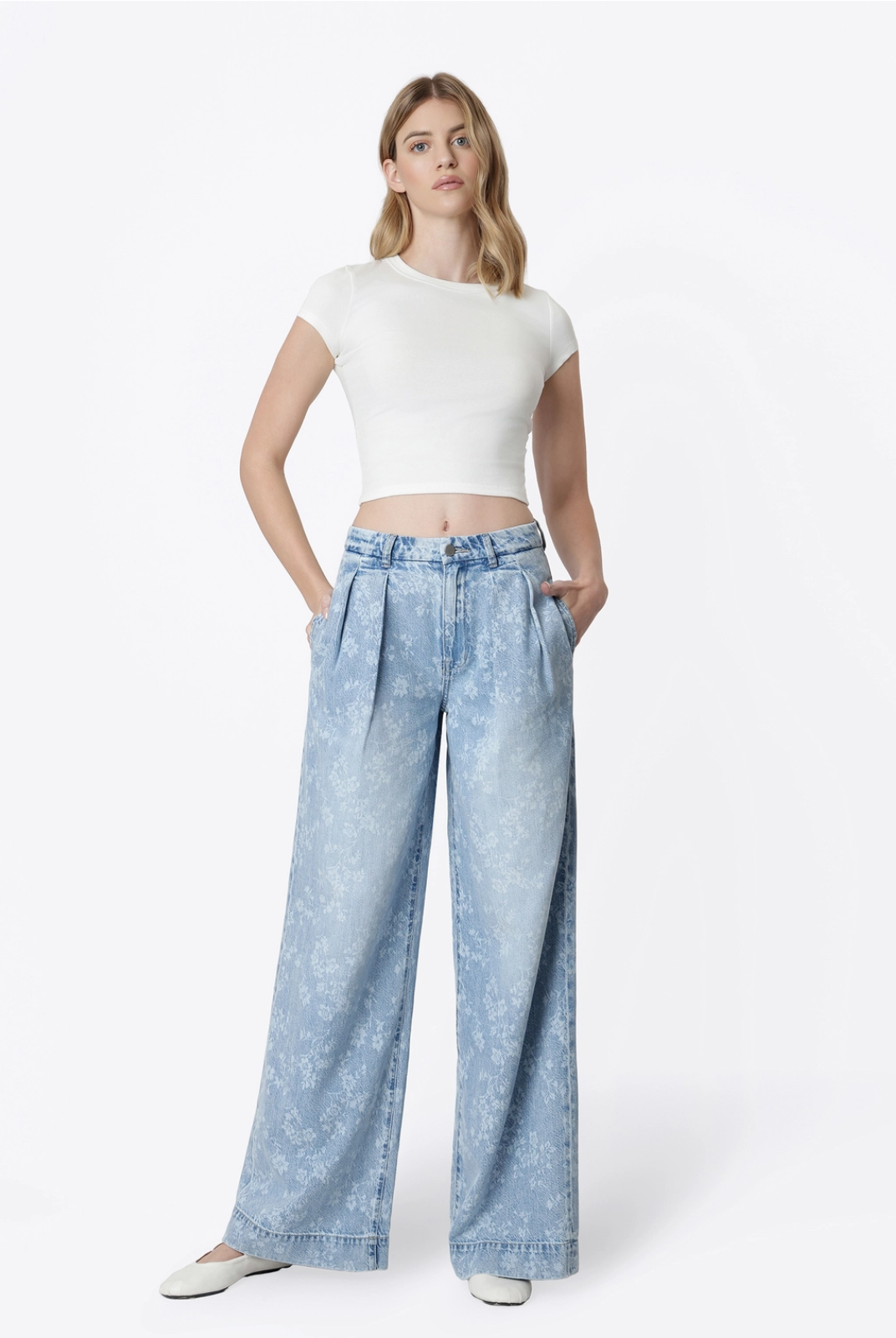 Model wearing Flying Monkey Hera Super High Rise Wide Leg Jeans in a light blue floral print wash, featuring a super high-rise waist, pleated front, relaxed wide-leg silhouette, subtle allover floral pattern, side pockets, and a full-length fit. Styled with a fitted white cropped tee and white flats.