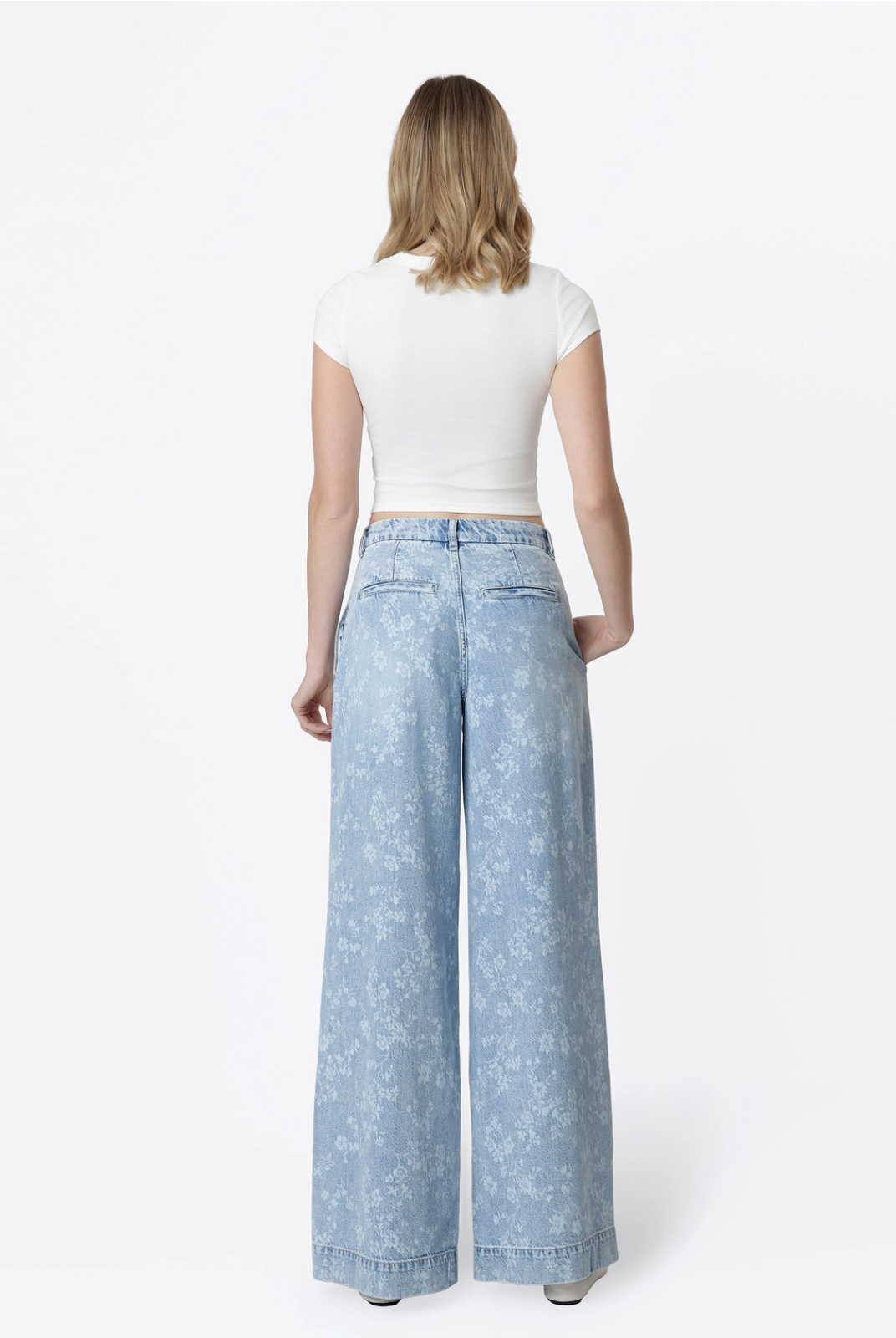 Model wearing Flying Monkey Hera Super High Rise Wide Leg Jeans in a light blue floral print wash, featuring a super high-rise waist, pleated front, relaxed wide-leg silhouette, subtle allover floral pattern, side pockets, and a full-length fit. Styled with a fitted white cropped tee and white flats.