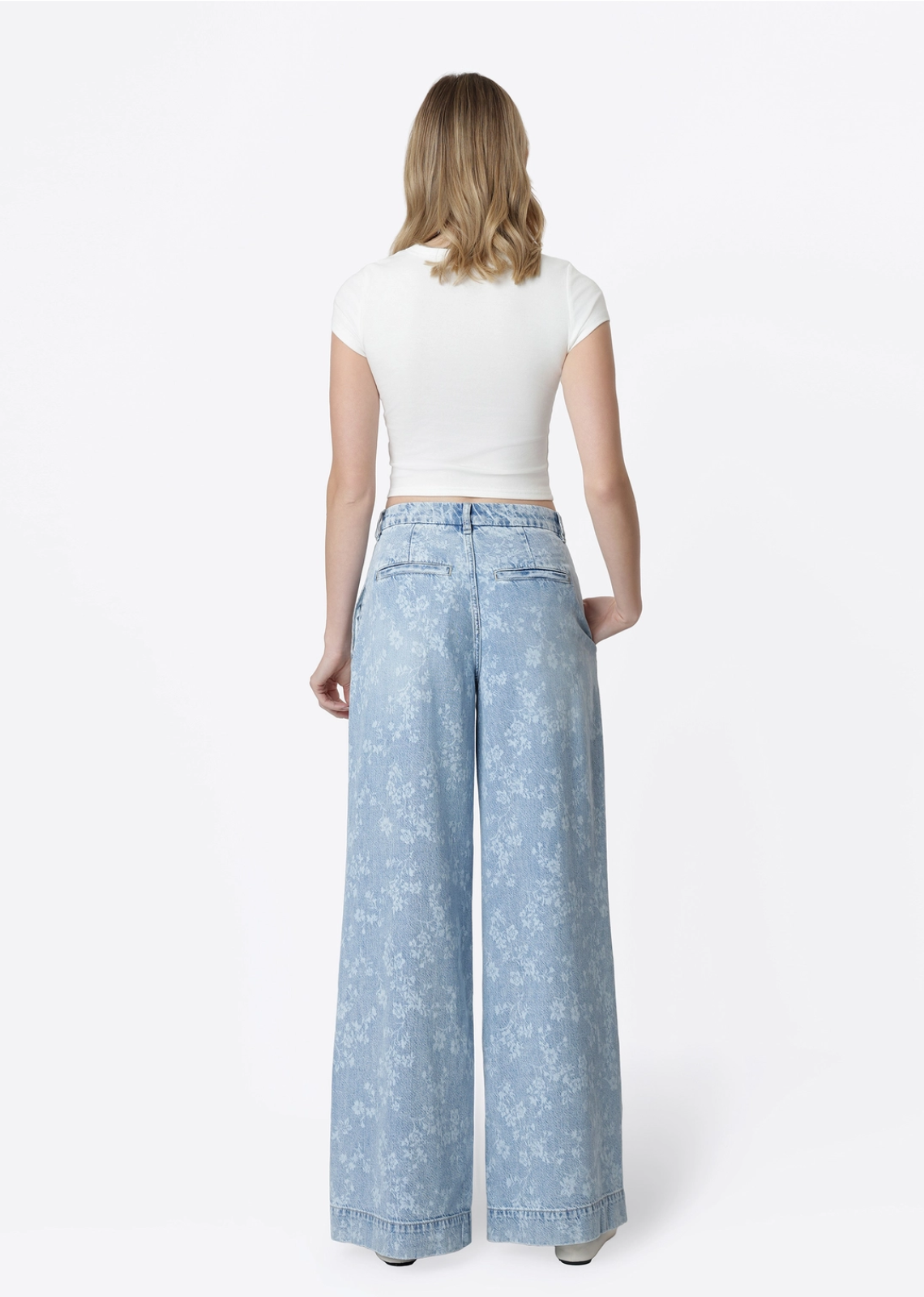 Model wearing Flying Monkey Hera Super High Rise Wide Leg Jeans in a light blue floral print wash, featuring a super high-rise waist, pleated front, relaxed wide-leg silhouette, subtle allover floral pattern, side pockets, and a full-length fit. Styled with a fitted white cropped tee and white flats.