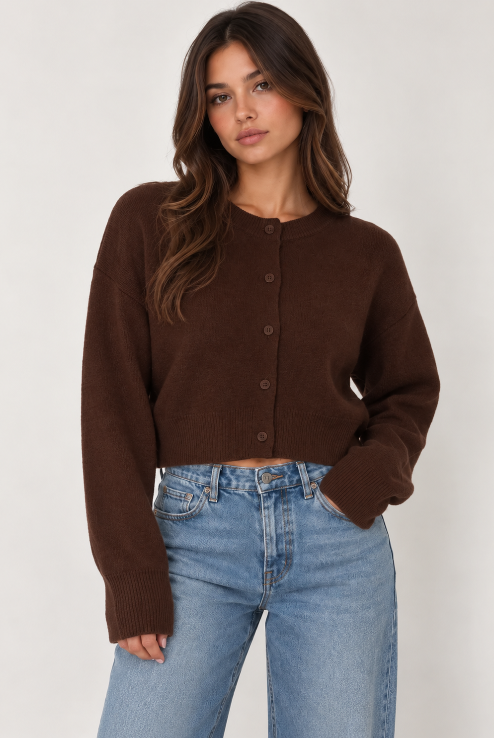 Model wearing a chocolate brown cropped knit cardigan featuring a classic crew neckline, button-down front, relaxed fit, dropped shoulders, long sleeves, and ribbed cuffs and hem. Crafted from soft, cozy knit fabric and styled with light-wash blue jeans for a casual, everyday look.