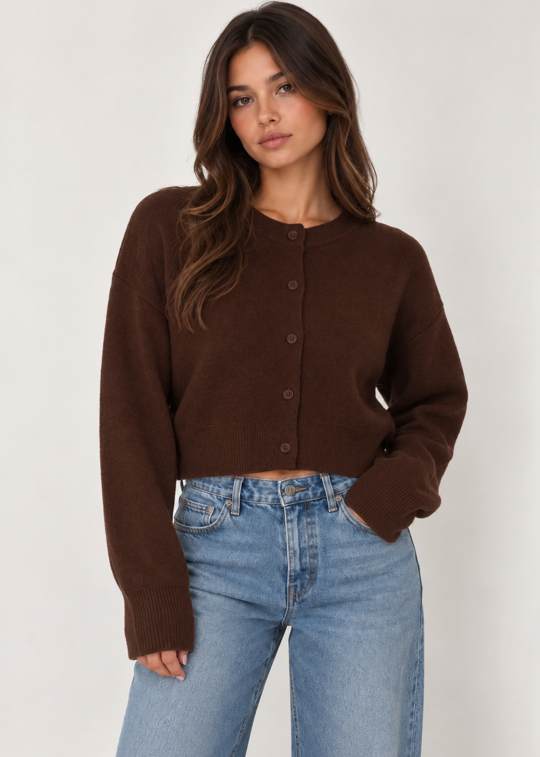 Model wearing a chocolate brown cropped knit cardigan featuring a classic crew neckline, button-down front, relaxed fit, dropped shoulders, long sleeves, and ribbed cuffs and hem. Crafted from soft, cozy knit fabric and styled with light-wash blue jeans for a casual, everyday look.
