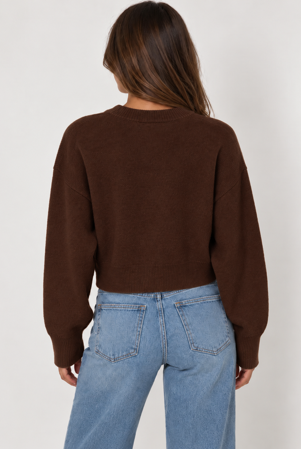 Model wearing a chocolate brown cropped knit cardigan featuring a classic crew neckline, button-down front, relaxed fit, dropped shoulders, long sleeves, and ribbed cuffs and hem. Crafted from soft, cozy knit fabric and styled with light-wash blue jeans for a casual, everyday look.