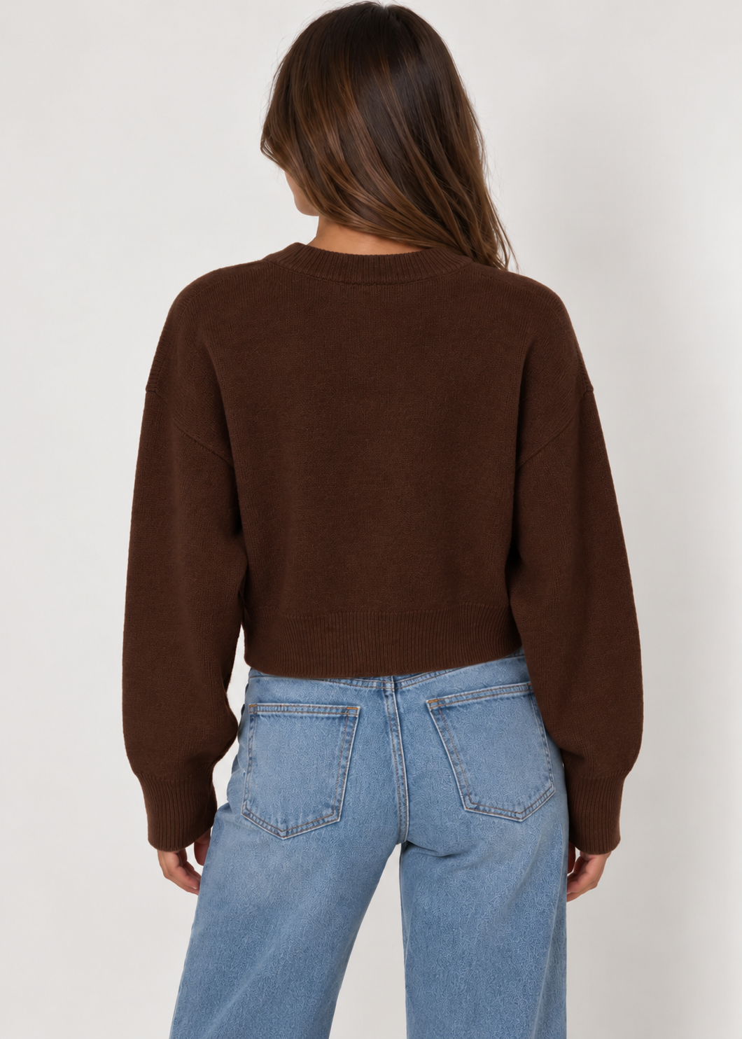 Model wearing a chocolate brown cropped knit cardigan featuring a classic crew neckline, button-down front, relaxed fit, dropped shoulders, long sleeves, and ribbed cuffs and hem. Crafted from soft, cozy knit fabric and styled with light-wash blue jeans for a casual, everyday look.
