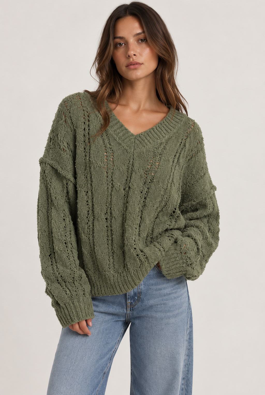Model wearing an oversized textured cable-knit V-neck sweater in olive, featuring an open-knit design, dropped shoulders, long sleeves, and ribbed trim, styled with light-wash denim.