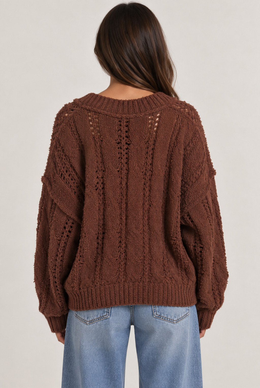 Model wearing an oversized textured cable-knit V-neck sweater in brown, featuring an open-knit design, dropped shoulders, long sleeves, and ribbed trim, styled with light-wash denim.