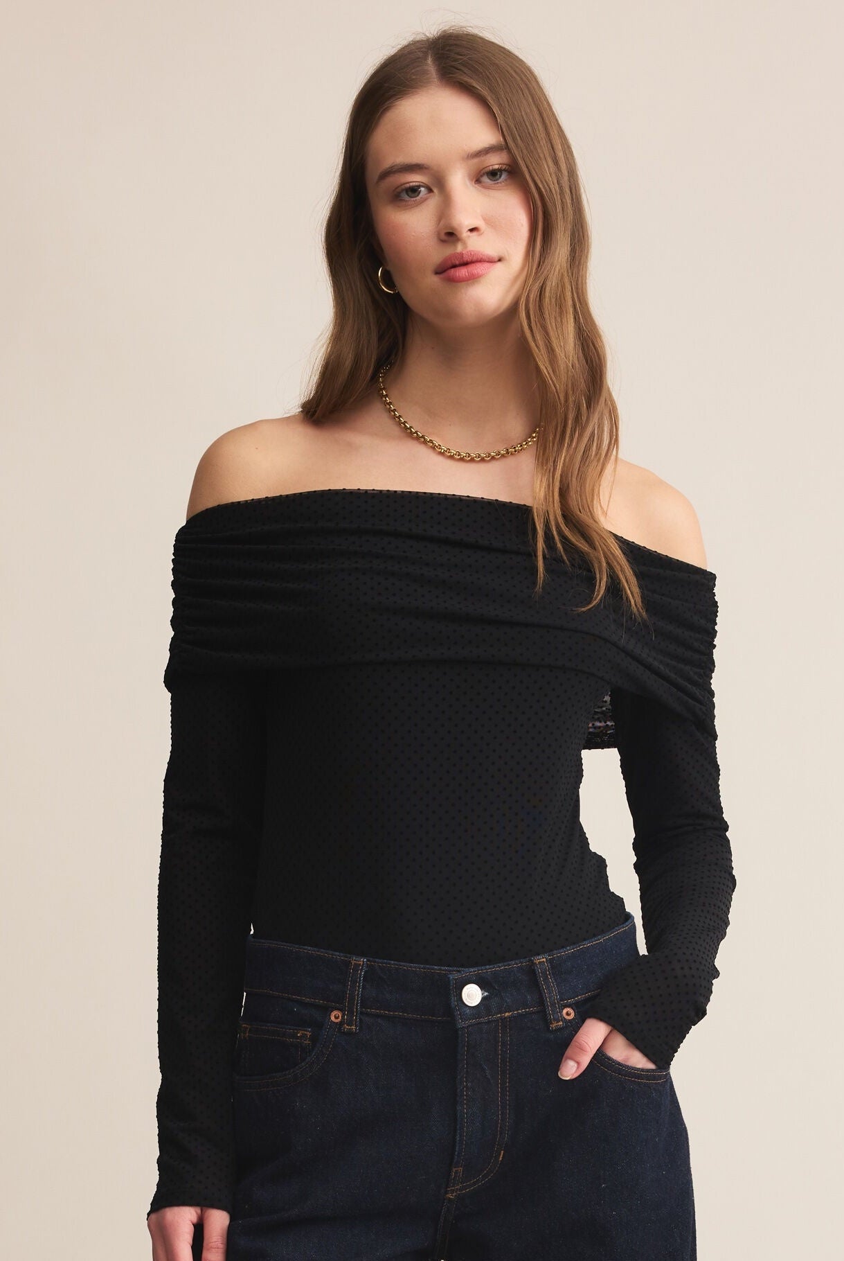Woman wearing a black off-shoulder top and dark jeans on a beige background