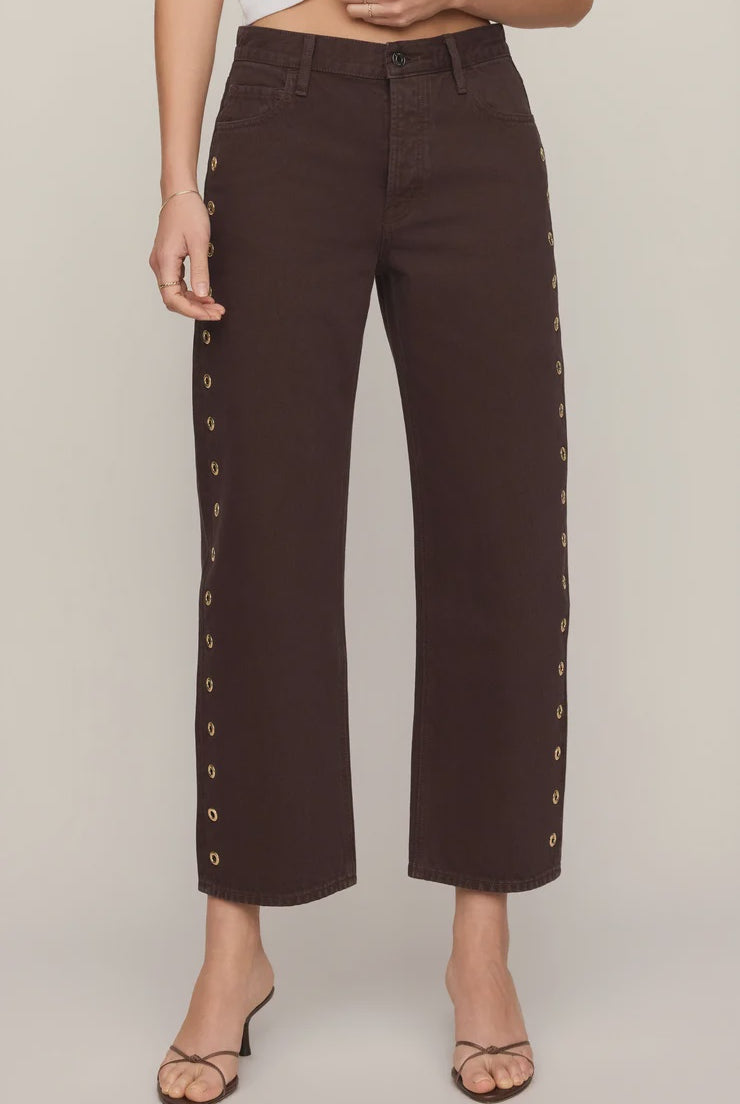 Rich brown mid-rise denim jeans featuring a relaxed bowed-leg silhouette, ankle-length hem, gold grommet details, and subtle side openings.