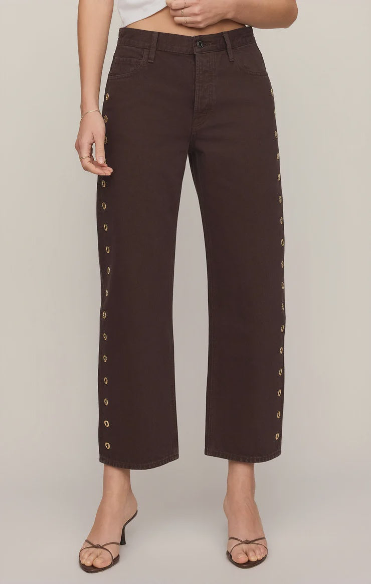 Rich brown mid-rise denim jeans featuring a relaxed bowed-leg silhouette, ankle-length hem, gold grommet details, and subtle side openings.