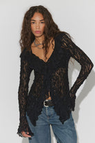 Textured lace cardigan in black, featuring a fitted longline silhouette, V-neckline, button-front closure, fitted long sleeves, and cascading ruffle hem detailing.
