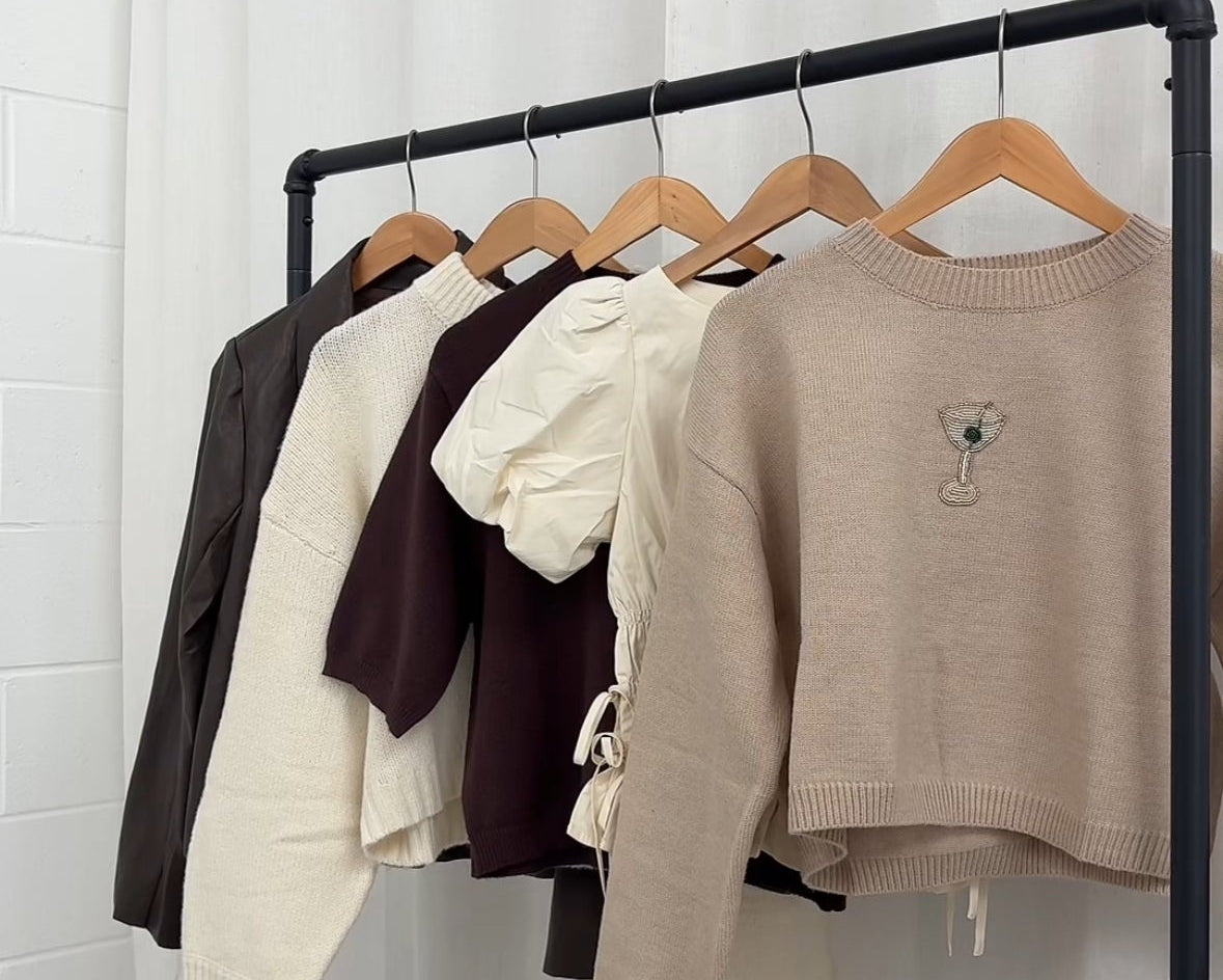 Clothing rack with various sweaters on hangers against a white curtain background