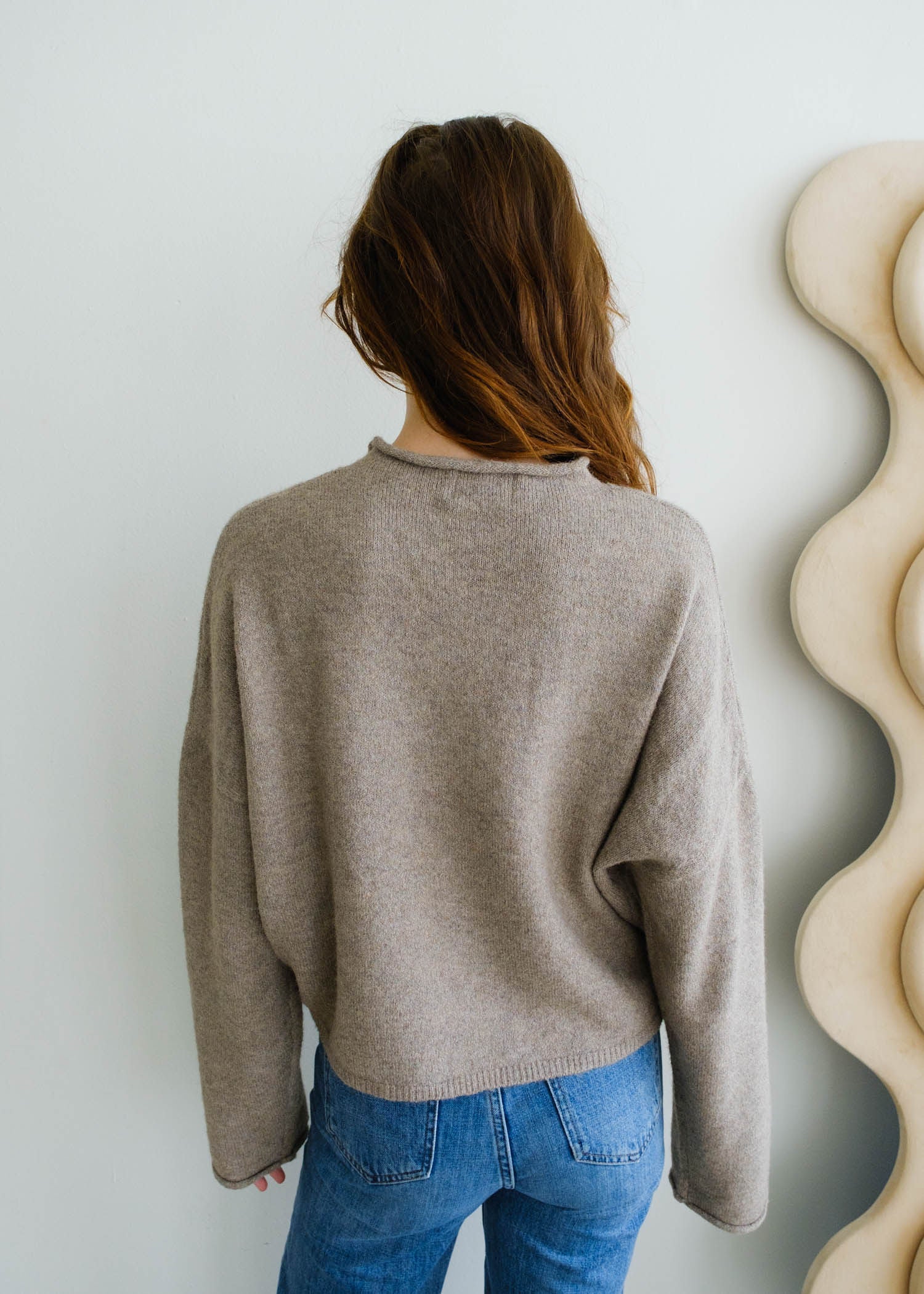Person wearing a beige sweater and blue jeans against a white wall with decorative elements.