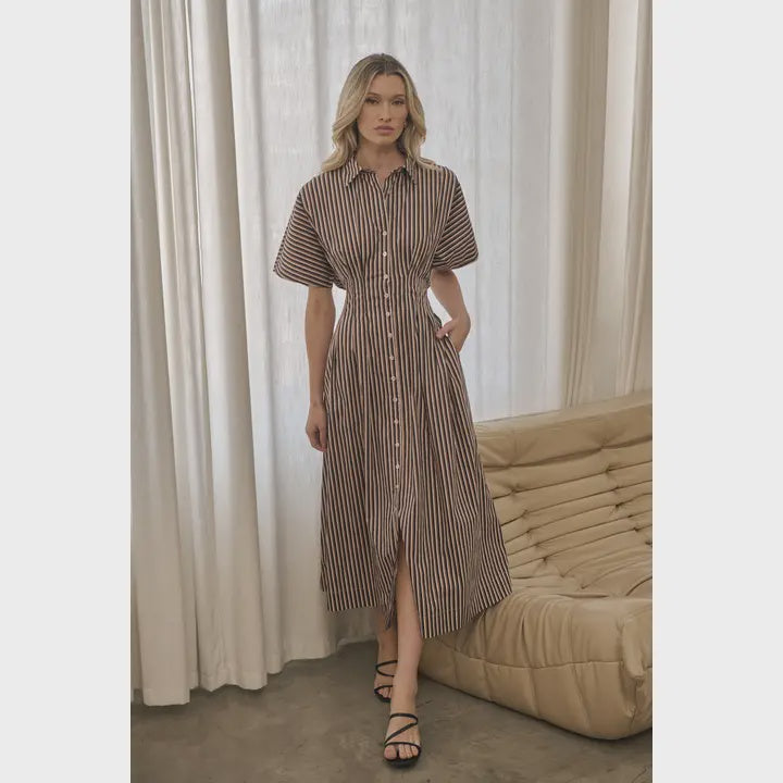 Model wearing The Emerson Pleated Midi Dress, a brown and ivory striped button-front midi dress featuring a collared neckline, short sleeves, a fitted waist, pleated skirt, and side pockets, styled with black strappy sandals.