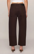 Rich brown mid-rise denim jeans featuring a relaxed bowed-leg silhouette, ankle-length hem, gold grommet details, and subtle side openings.
