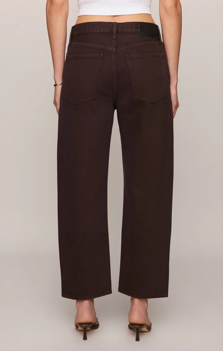 Rich brown mid-rise denim jeans featuring a relaxed bowed-leg silhouette, ankle-length hem, gold grommet details, and subtle side openings.
