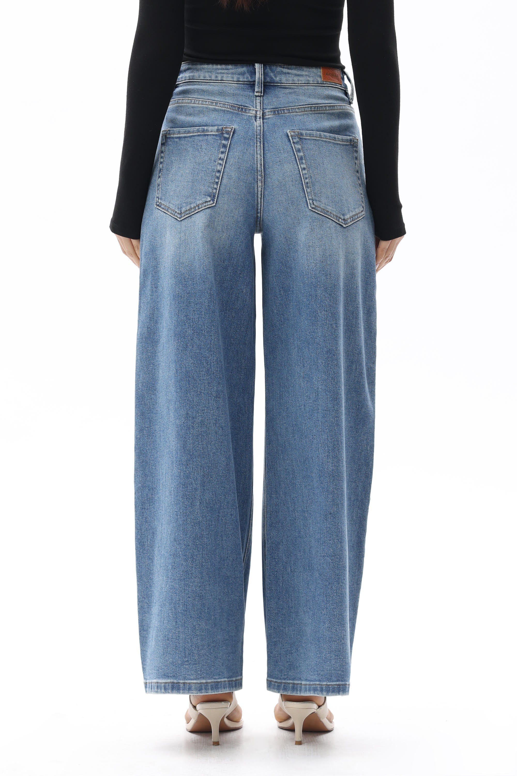 Model wearing BAYEAS Valentine High Rise Barrel Jeans in a medium blue wash, featuring a high-rise waist, relaxed barrel-leg silhouette, subtle fading, front panel seam detailing, classic five-pocket styling, and a full-length fit. Styled with a black long-sleeve top and nude thong sandals.
