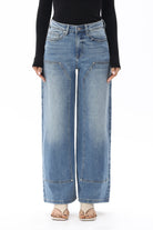 Model wearing BAYEAS Valentine High Rise Barrel Jeans in a medium blue wash, featuring a high-rise waist, relaxed barrel-leg silhouette, subtle fading, front panel seam detailing, classic five-pocket styling, and a full-length fit. Styled with a black long-sleeve top and nude thong sandals.
