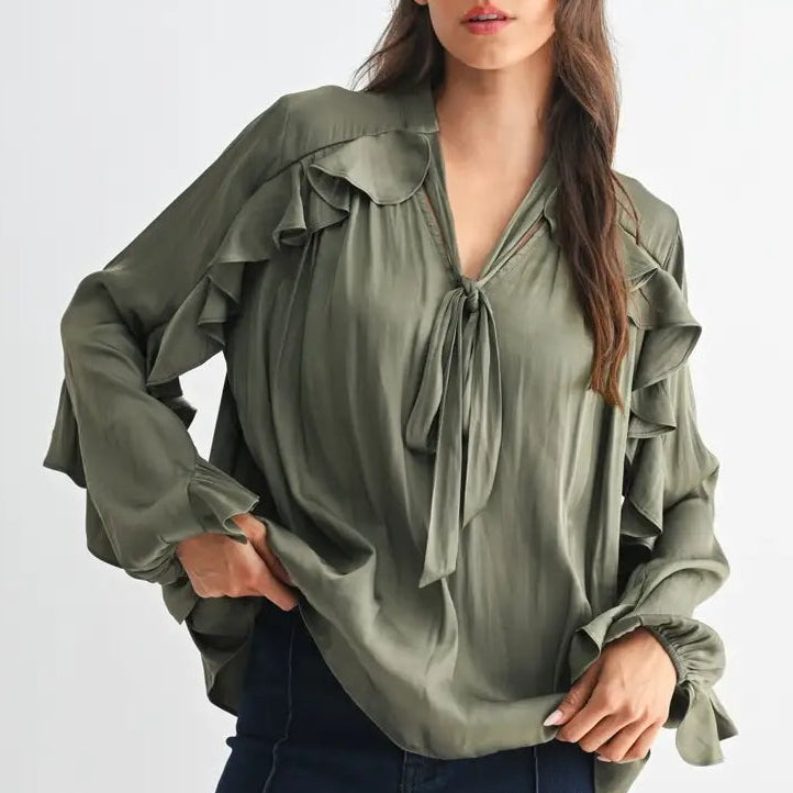 A woman modeling a sage green long-sleeve blouse with ruffle details and a tie-neck, paired with dark blue jeans, against a light gray studio background.
