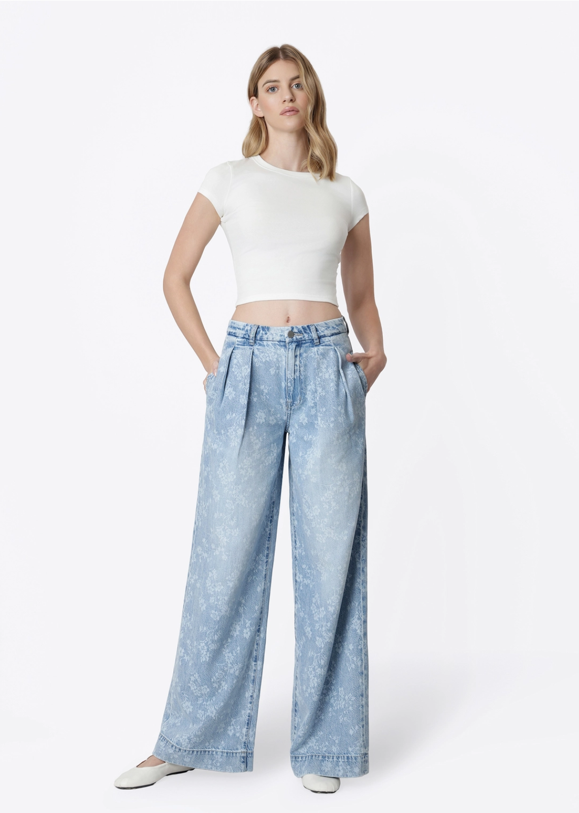 Model wearing Flying Monkey Hera Super High Rise Wide Leg Jeans in a light blue floral print wash, featuring a super high-rise waist, pleated front, relaxed wide-leg silhouette, subtle allover floral pattern, side pockets, and a full-length fit. Styled with a fitted white cropped tee and white flats.
