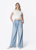 Model wearing Flying Monkey Hera Super High Rise Wide Leg Jeans in a light blue floral print wash, featuring a super high-rise waist, pleated front, relaxed wide-leg silhouette, subtle allover floral pattern, side pockets, and a full-length fit. Styled with a fitted white cropped tee and white flats.
