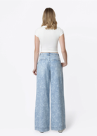 Model wearing Flying Monkey Hera Super High Rise Wide Leg Jeans in a light blue floral print wash, featuring a super high-rise waist, pleated front, relaxed wide-leg silhouette, subtle allover floral pattern, side pockets, and a full-length fit. Styled with a fitted white cropped tee and white flats.
