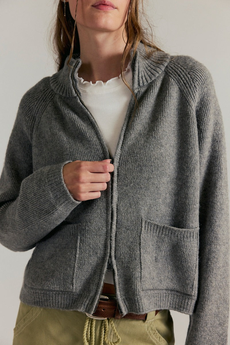 Women's ribbed knit zip-front sweater in cool grey with a high turtleneck collar, raglan sleeves, and front patch pockets.