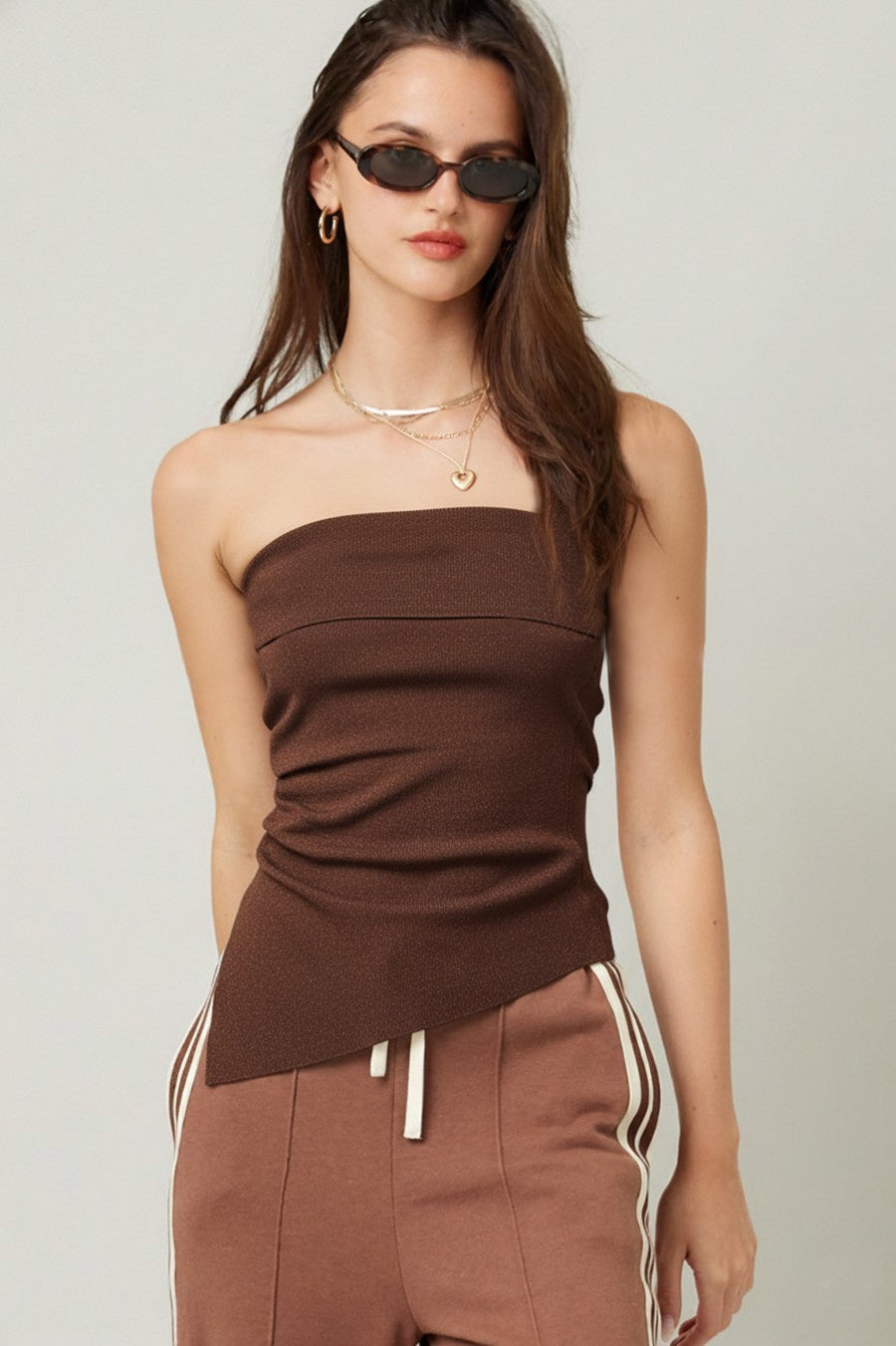 Model wearing a strapless chocolate brown knit tube top with subtle ruching through the waist and an asymmetrical hem. 