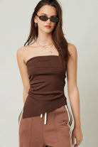 Model wearing a strapless chocolate brown knit tube top with subtle ruching through the waist and an asymmetrical hem. 