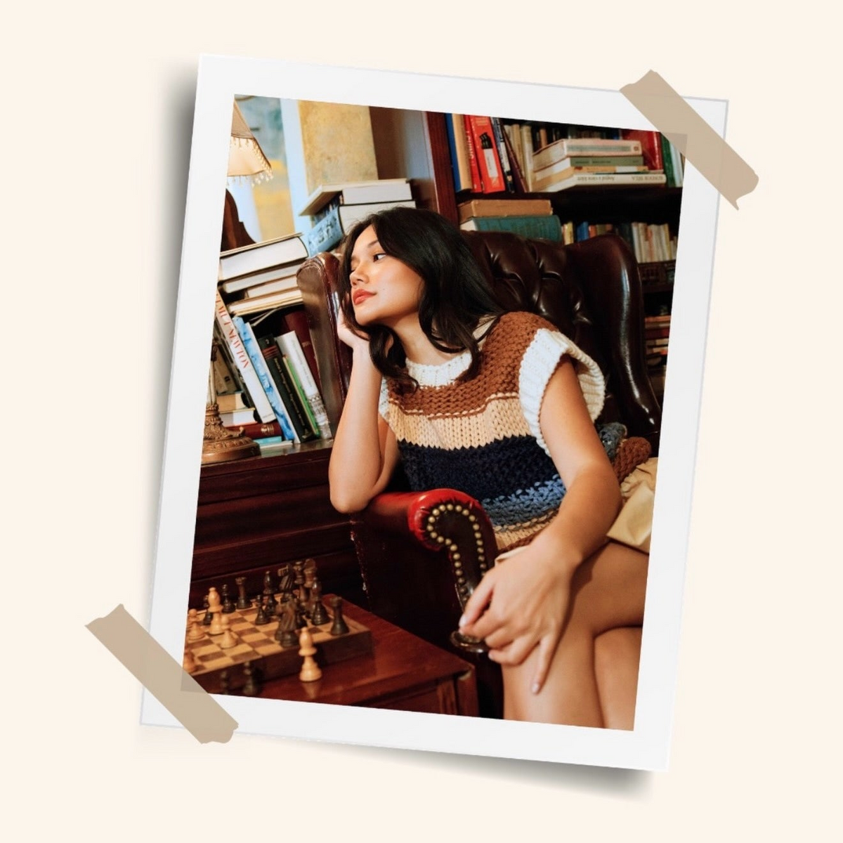 A Polaroid-style photo print showing a woman sitting in a leather armchair in a book-filled room, wearing a crocheted sleeveless top. The print is displayed on a light beige background with two pieces of tape at the corners.