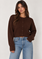 Model wearing a chocolate brown cropped knit cardigan featuring a classic crew neckline, button-down front, relaxed fit, dropped shoulders, long sleeves, and ribbed cuffs and hem. Crafted from soft, cozy knit fabric and styled with light-wash blue jeans for a casual, everyday look.