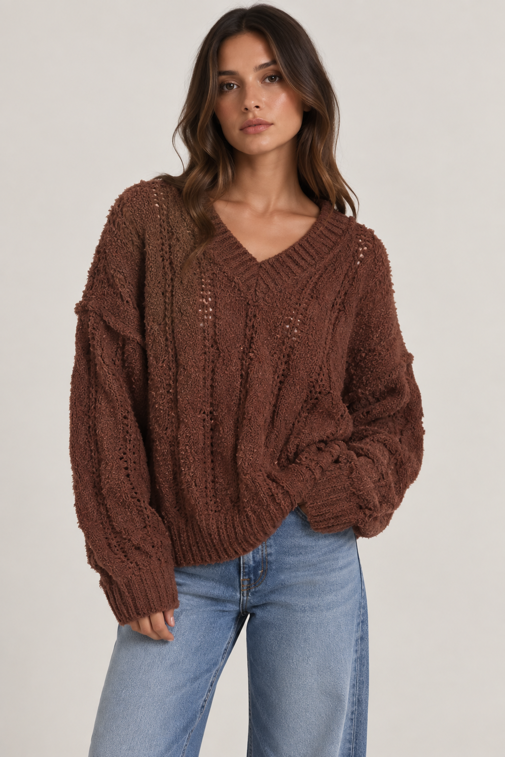 Model wearing an oversized textured cable-knit V-neck sweater in brown, featuring an open-knit design, dropped shoulders, long sleeves, and ribbed trim, styled with light-wash denim.