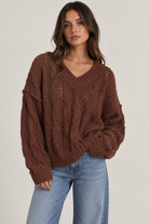 Model wearing an oversized textured cable-knit V-neck sweater in brown, featuring an open-knit design, dropped shoulders, long sleeves, and ribbed trim, styled with light-wash denim.