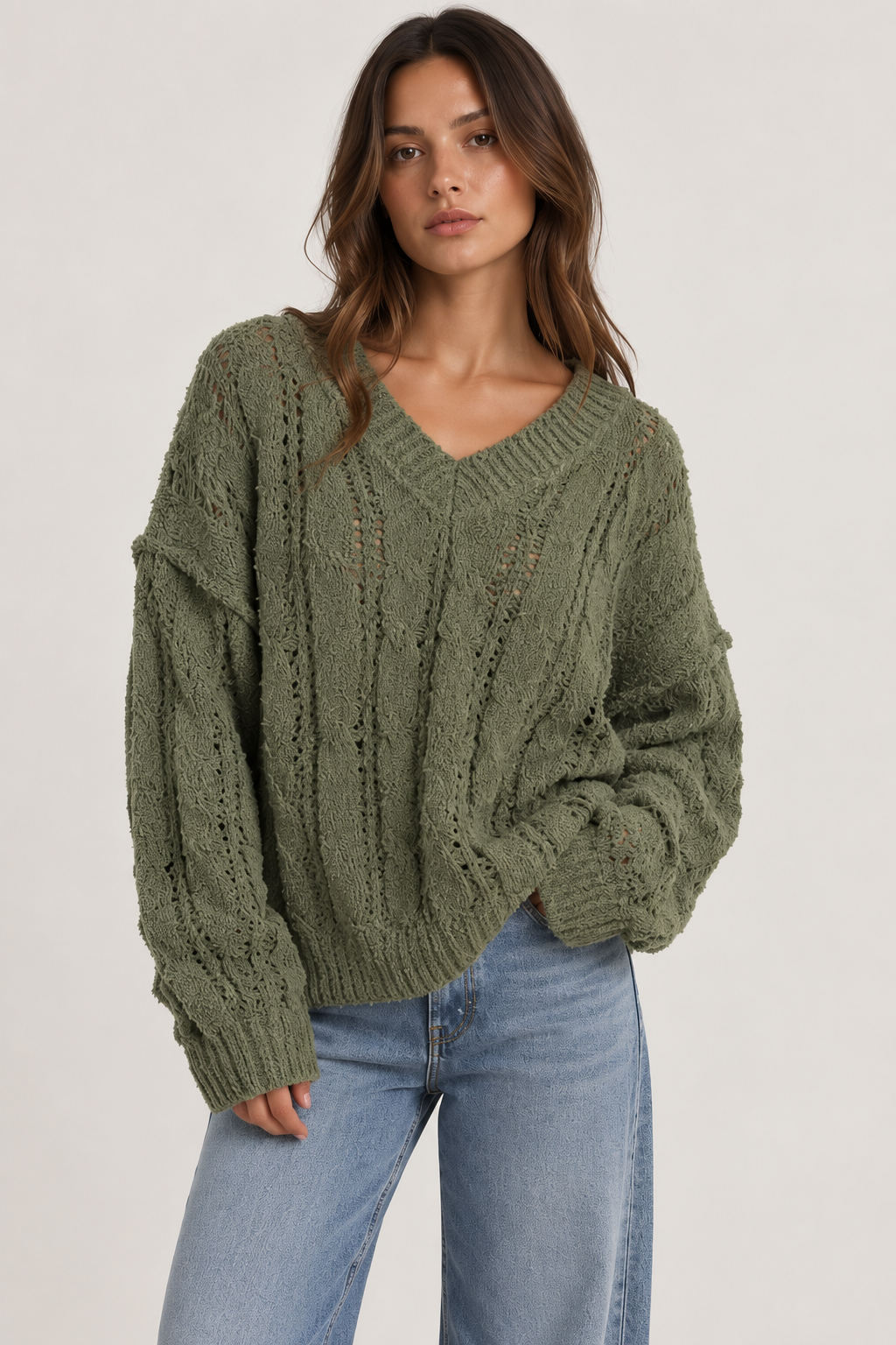 Model wearing an oversized textured cable-knit V-neck sweater in olive, featuring an open-knit design, dropped shoulders, long sleeves, and ribbed trim, styled with light-wash denim.